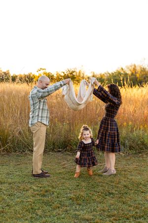 A family plays together in a golden field at sunset, lifting a blanket in the air while their child stands between them.