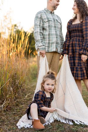 Family wearing coordinated plaid outfits poses together with white blanket in golden autumn field at sunset.