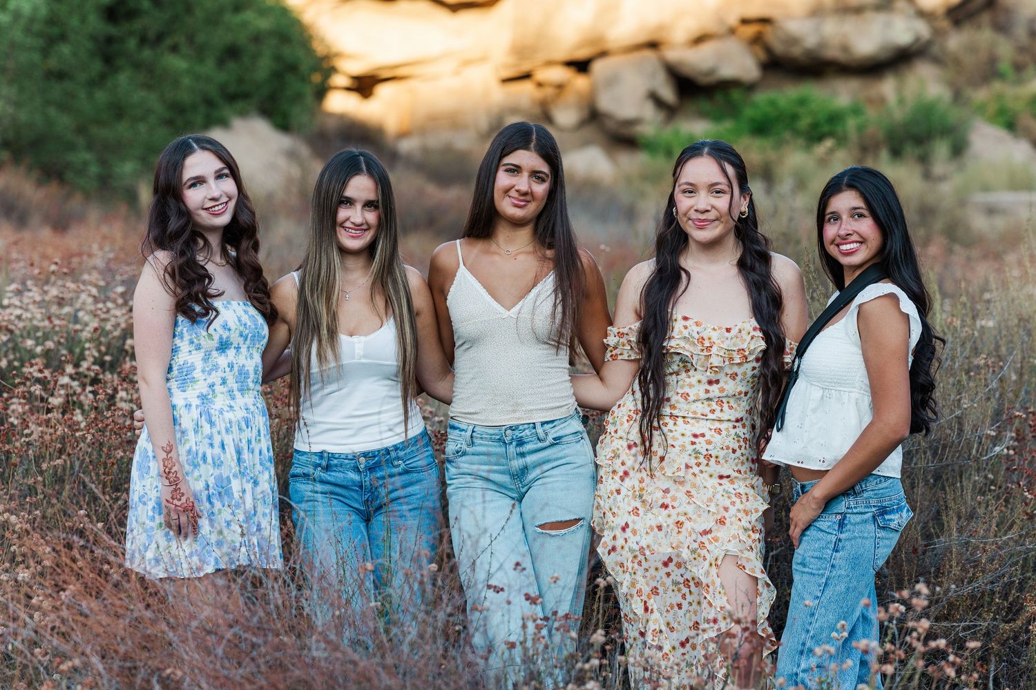 Five friends in casual denim and white outfits pose together outdoors in a natural setting.