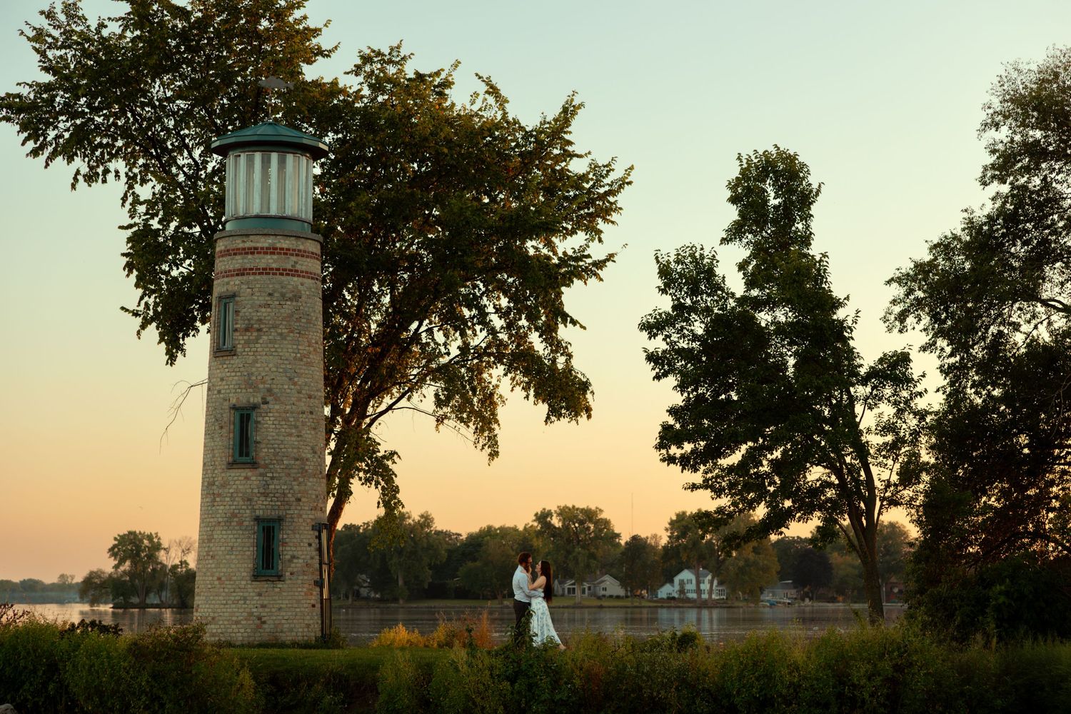 Historic lighthouse stands tall against a golden sunset sky with silhouetted trees framing the peaceful coastal scene.