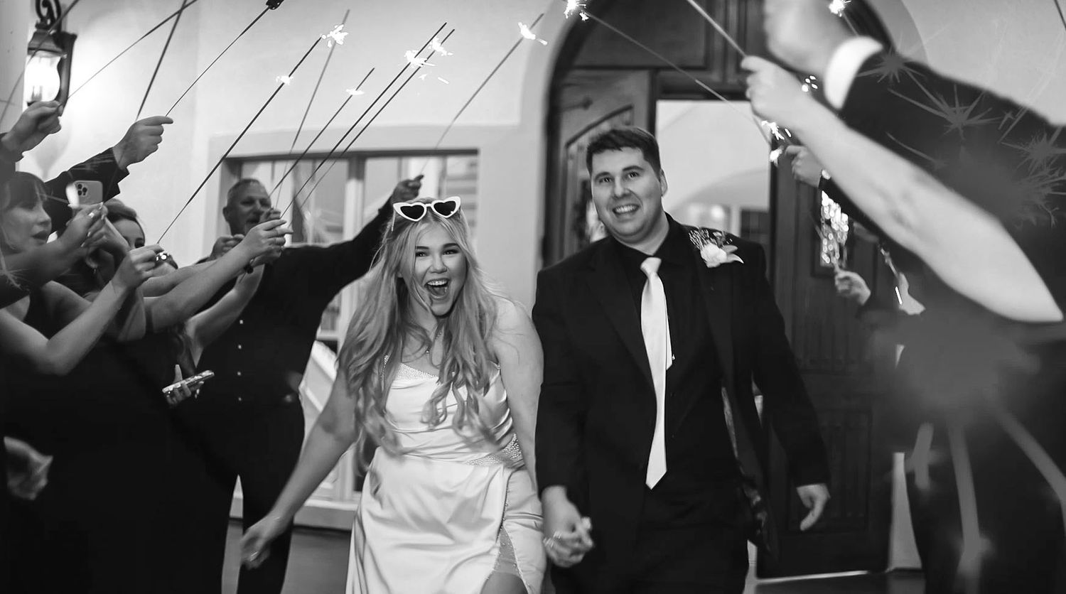 Excited bride and groom exit wedding reception through sparkler tunnel, bride laughing joyfully in black and white photo.