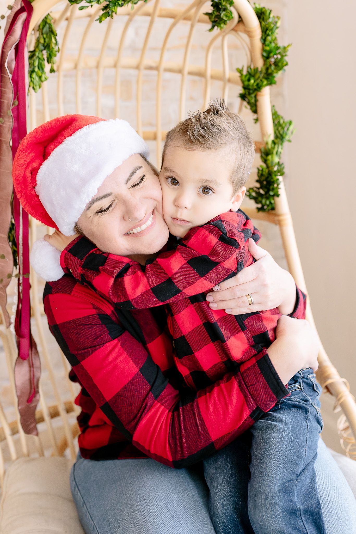 Festive holiday portrait with red buffalo plaid shirts and Santa hat against wicker chair and greenery backdrop.