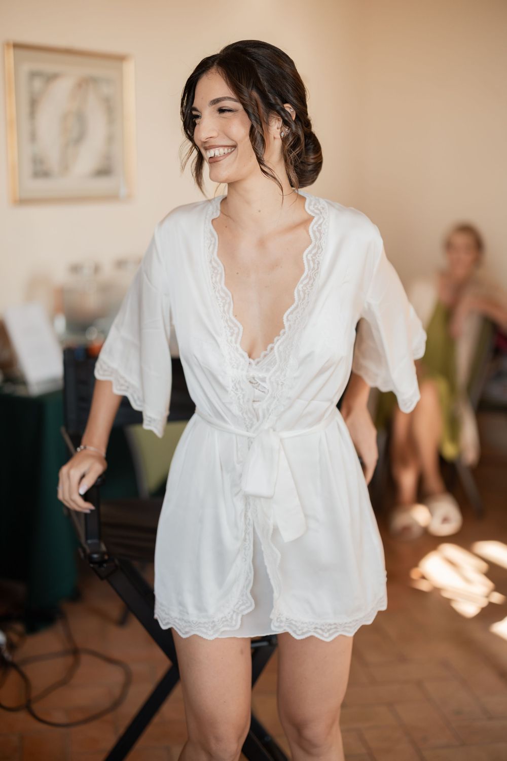 A white lace bridal robe with scalloped edges is worn during wedding preparations in a sunlit room.