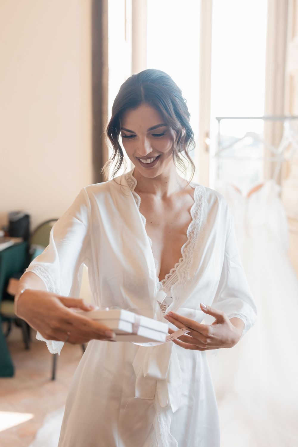 Bride in white robe opens letter near bright window during wedding day preparations.