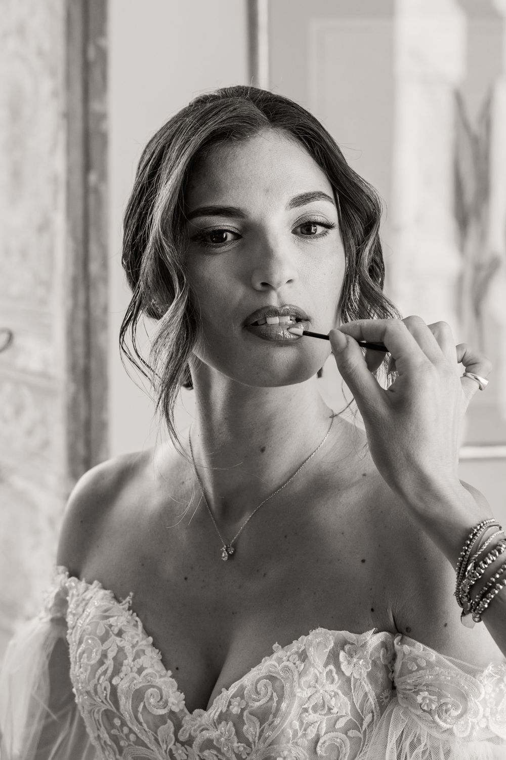 Black and white portrait of someone getting makeup applied while wearing an ornate lace wedding dress.