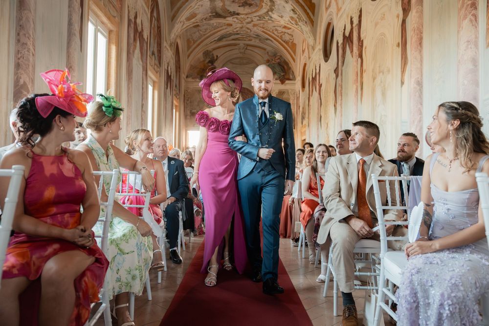 Wedding ceremony in an ornate hallway with guests seated in white chairs and colorful fascinators.