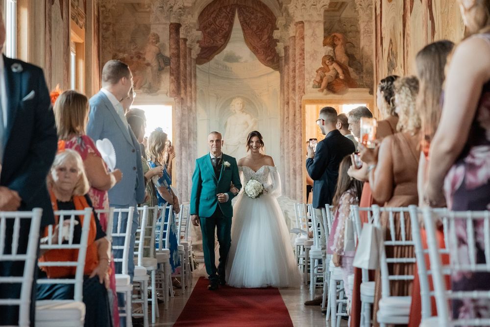 Wedding guests seated in white chairs during an elegant indoor ceremony with ornate architectural details.