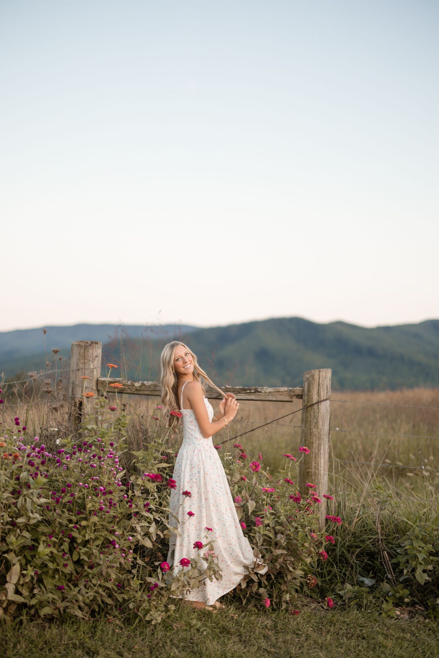 A white dress flows in the breeze against a backdrop of pink roses and mountain scenery at golden hour.