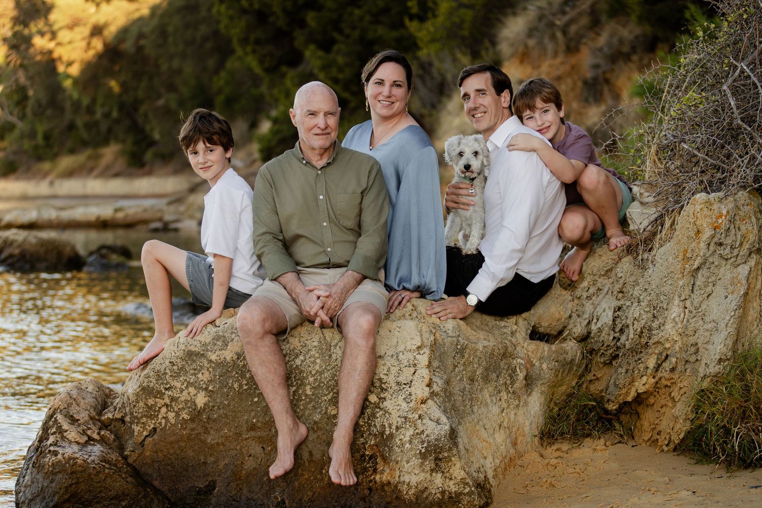 Family and their white dog pose together on large rocks at sunset by the beach during a photo session.
