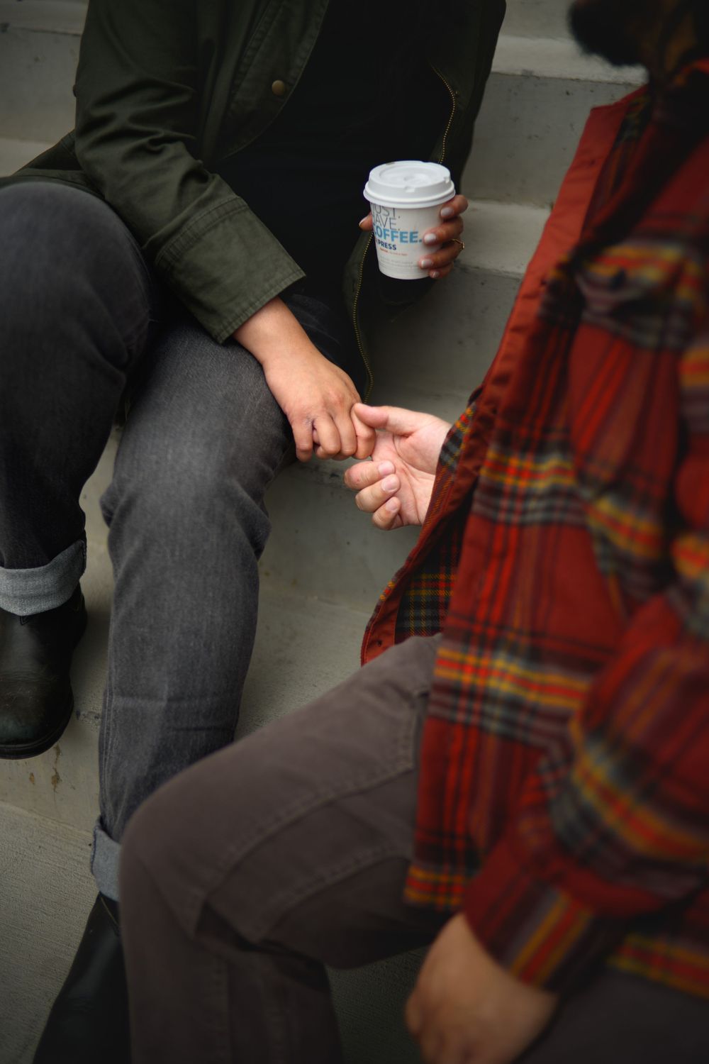Two people sitting close together on a bench with one person holding a coffee cup in casual attire.