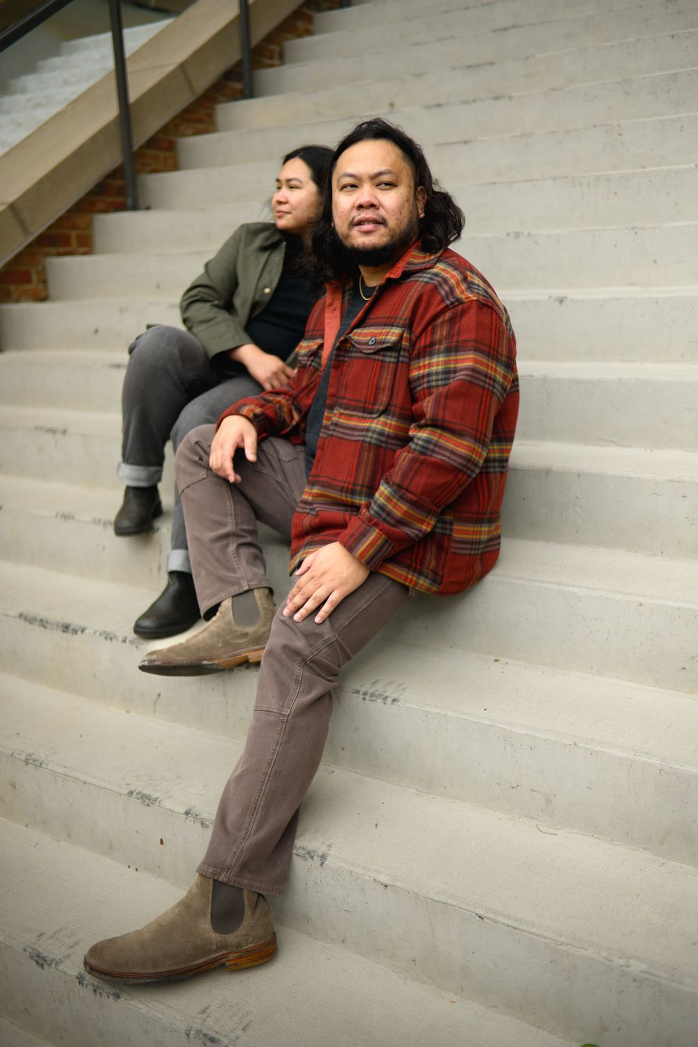 Two people sit casually on concrete steps wearing casual fall attire including plaid flannel and boots.
