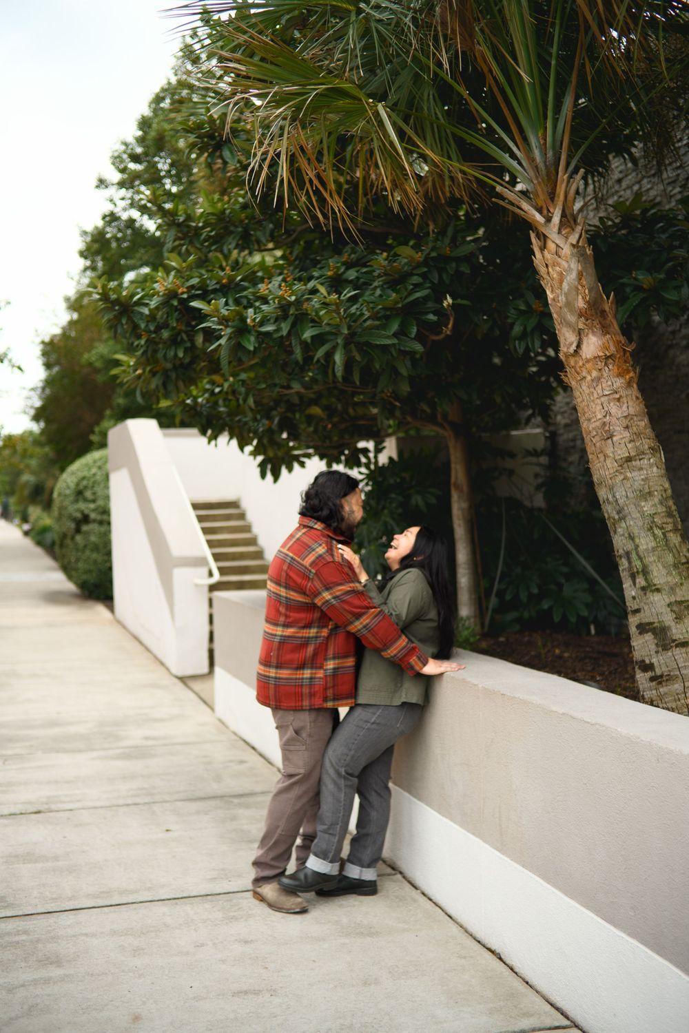 Person in plaid shirt and jeans leans against white wall while looking at mobile phone in outdoor setting.