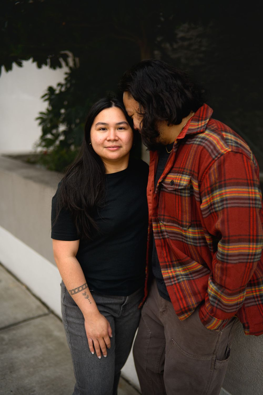 Two people in casual clothing, one wearing a black shirt and one in a plaid flannel, pose together outdoors.