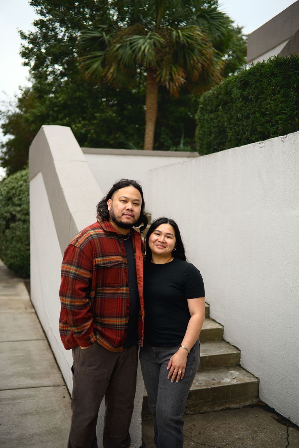 A couple poses together against a white wall in an outdoor setting wearing contrasting casual outfits.