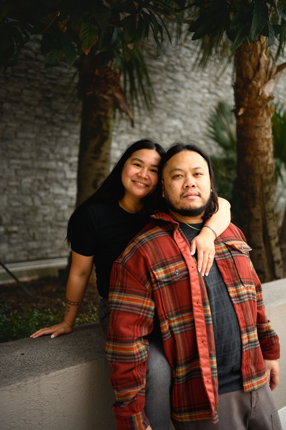 Two people in casual attire sharing a warm embrace and laughing together against a textured wall background.