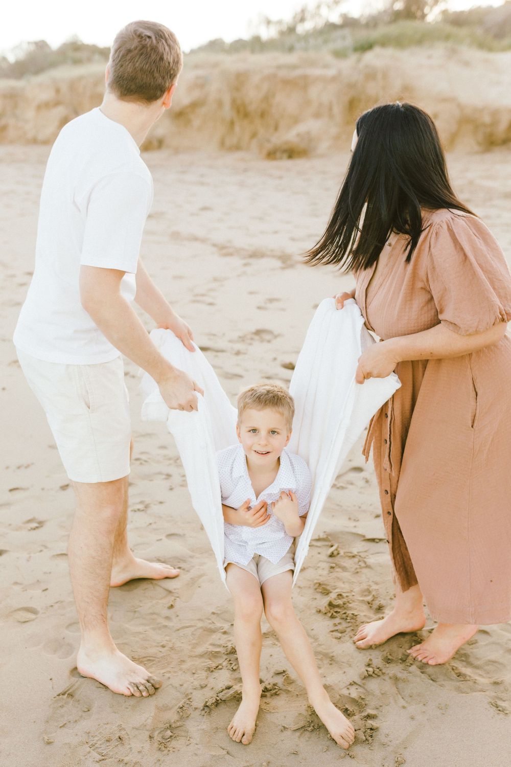 Family wearing white and tan outfits walking together on sandy beach during sunset photoshoot.