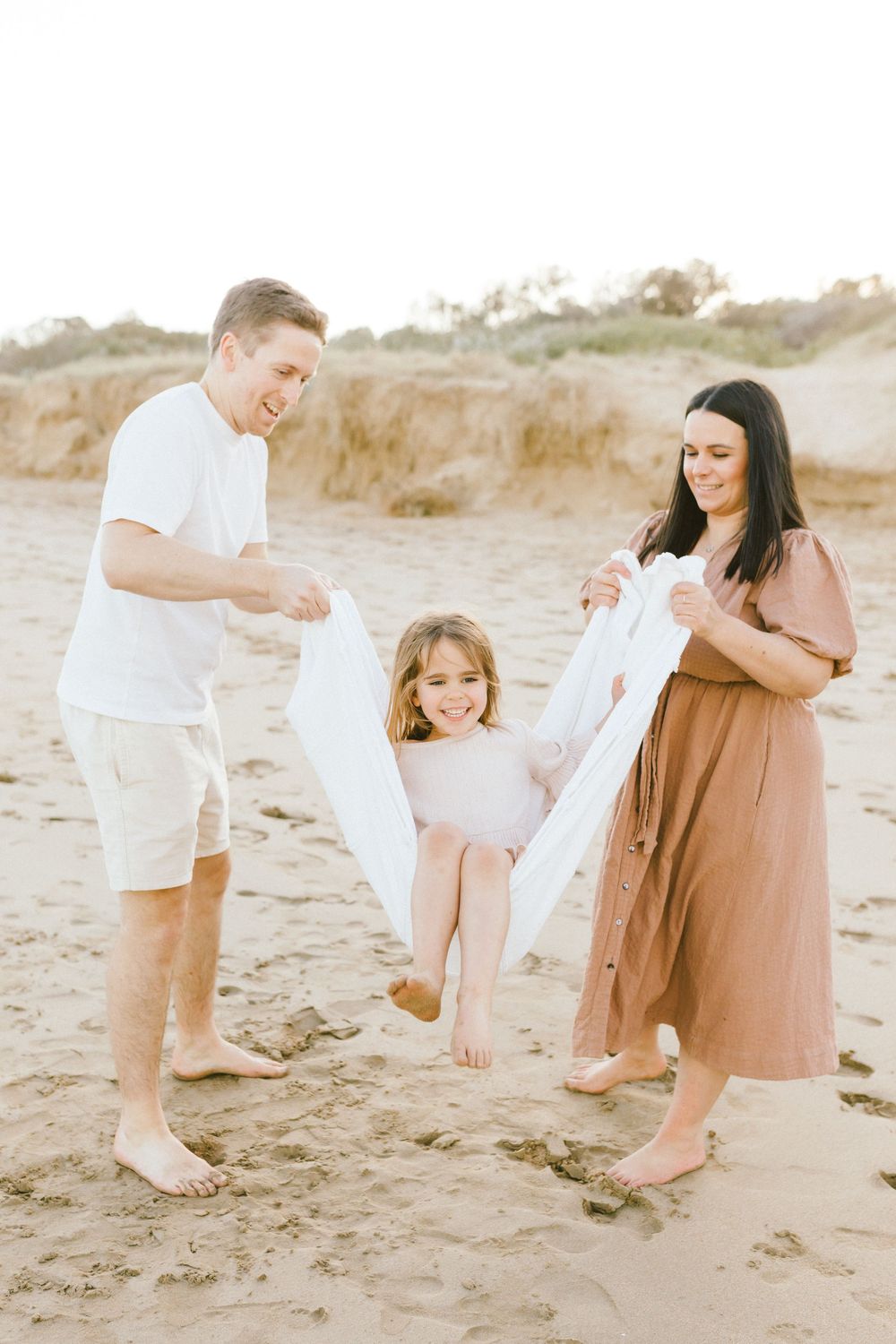 A family plays together on a sandy beach during a warm sunset photoshoot in neutral colored clothing.