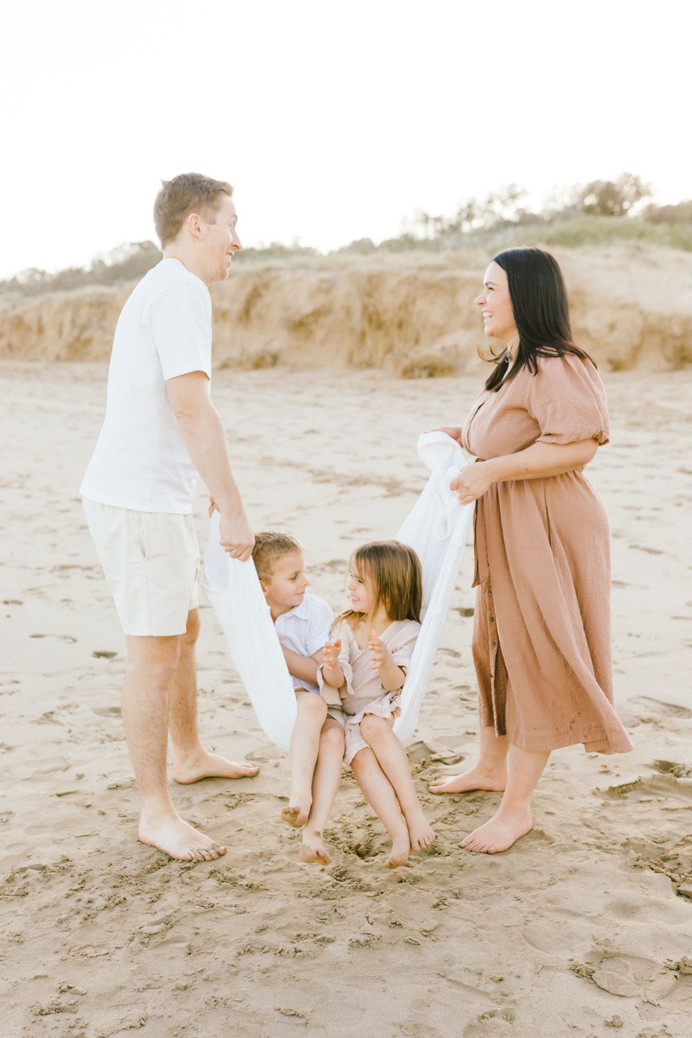 Family in light neutral clothing playing together on a sandy beach at sunset.