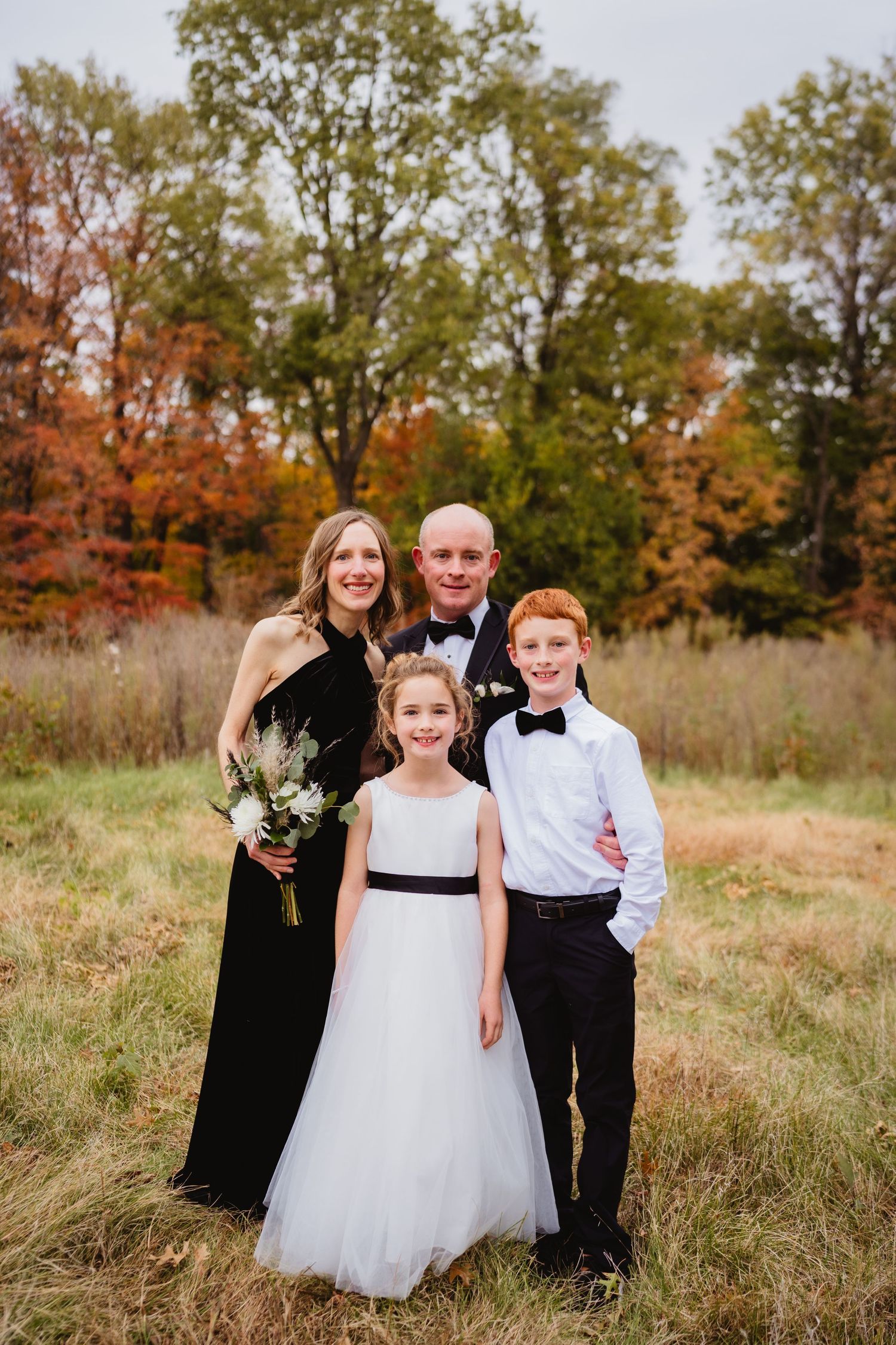 Wedding party poses together in autumn field with tall grass and colorful fall foliage in background.