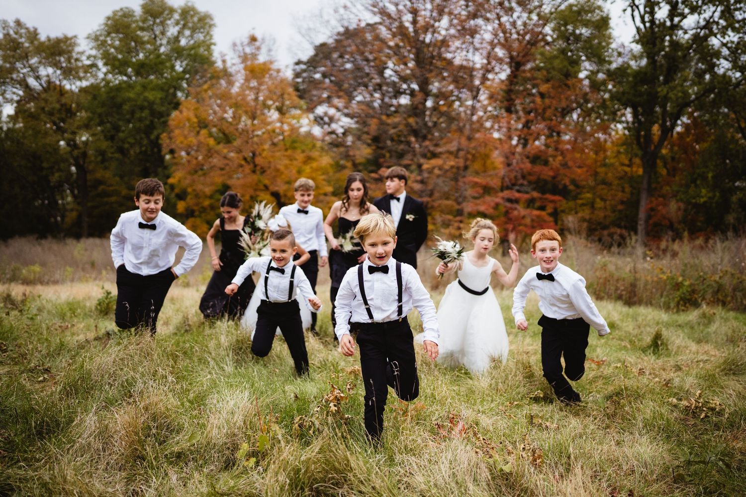 Wedding party runs and laughs together through a rustic autumn field with golden grass and fall foliage.