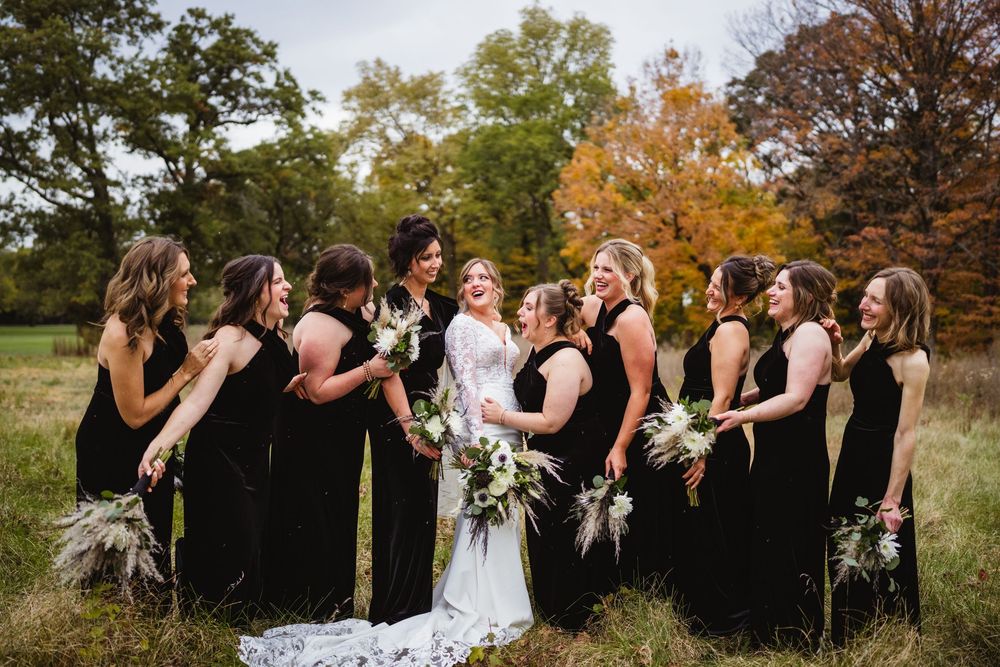 A wedding party wearing black dresses and formal attire gather outdoors in a field with autumn trees in the background.