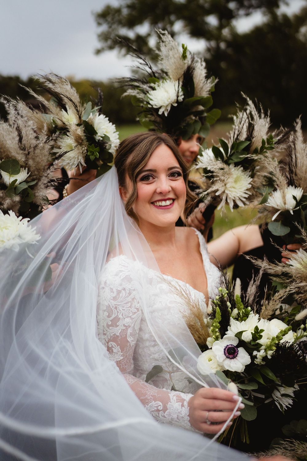 A bride in a white lace wedding dress smiles while holding a bouquet of white flowers and greenery.