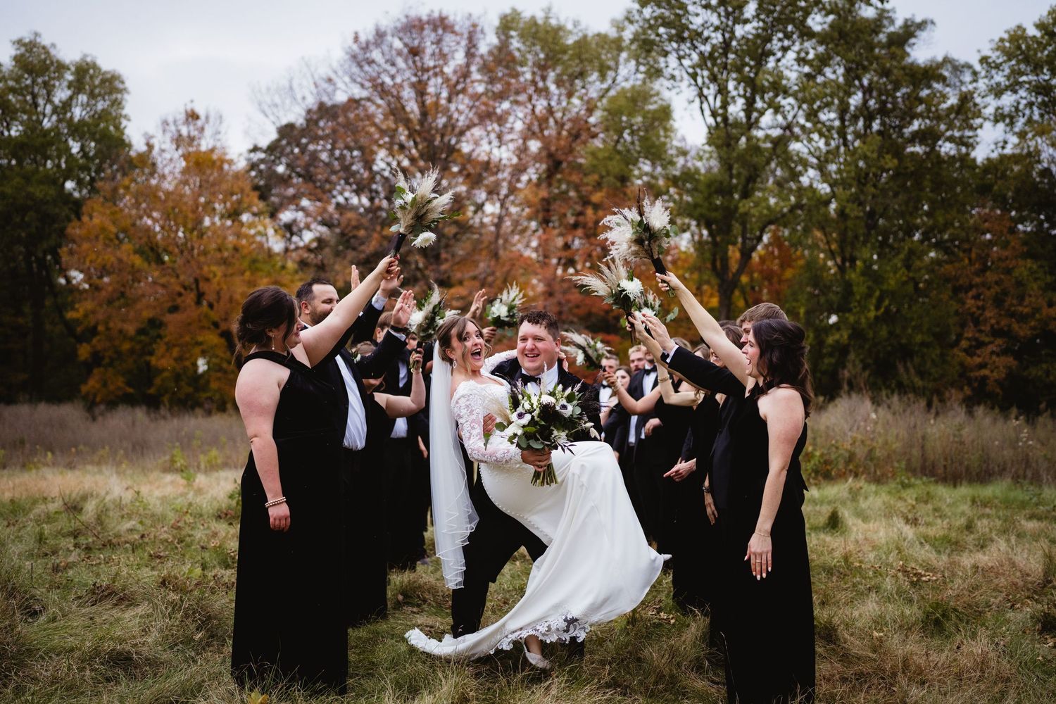 Wedding party celebrates outdoors with joyful jumping poses against autumn trees and natural landscape.