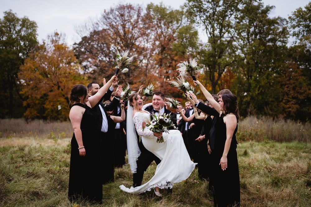 Wedding party celebrates outdoors with joyful jumping poses against autumn trees and natural landscape.