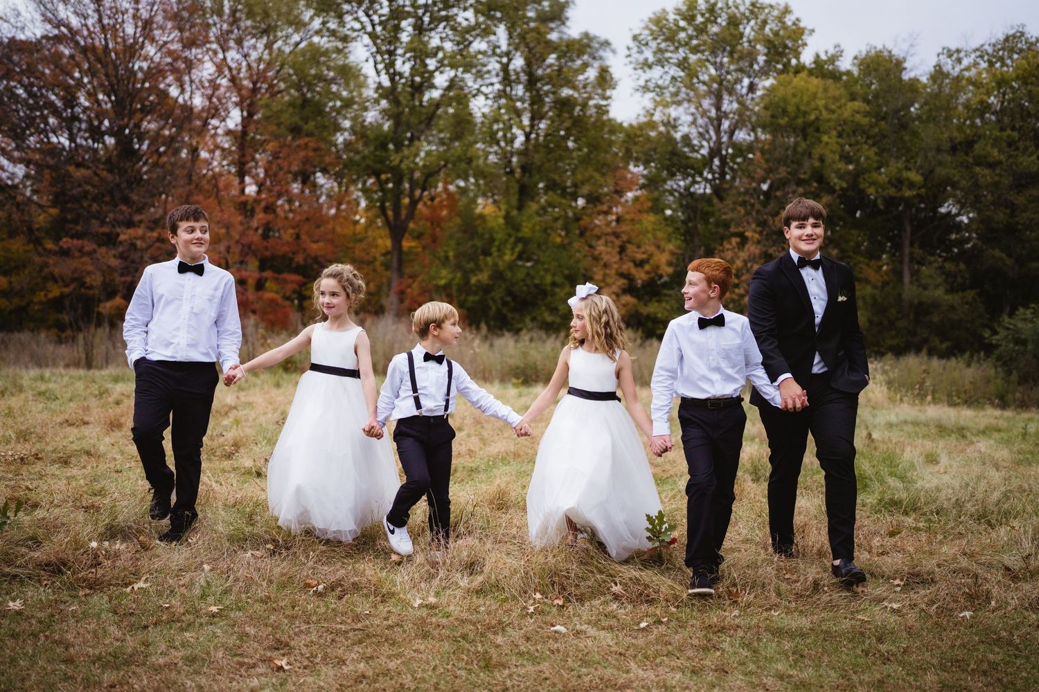 A wedding party walks together through a scenic autumn field with tall grass and trees in the background.