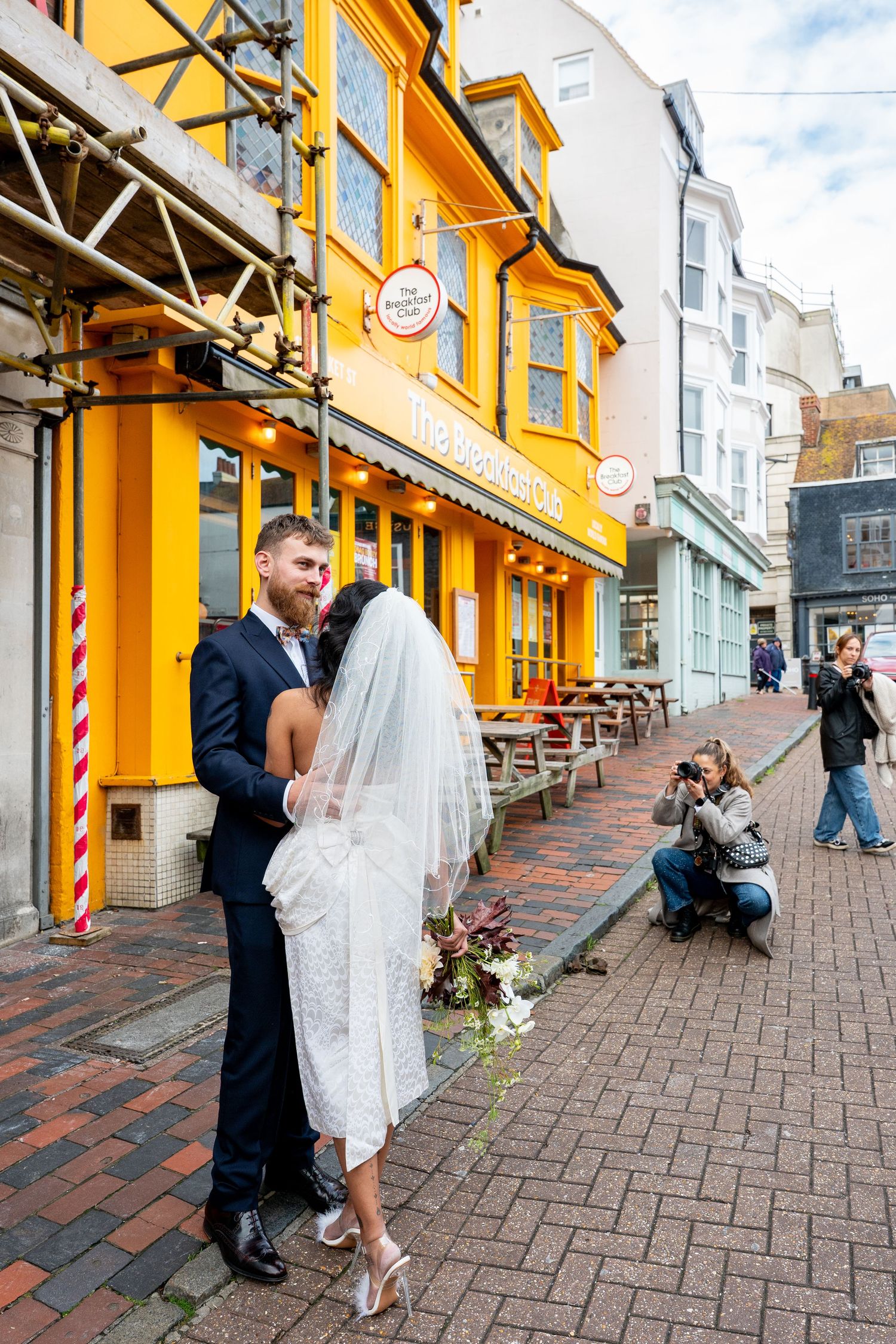 A couple shares a romantic moment outside a bright yellow storefront on a brick sidewalk in Brighton.