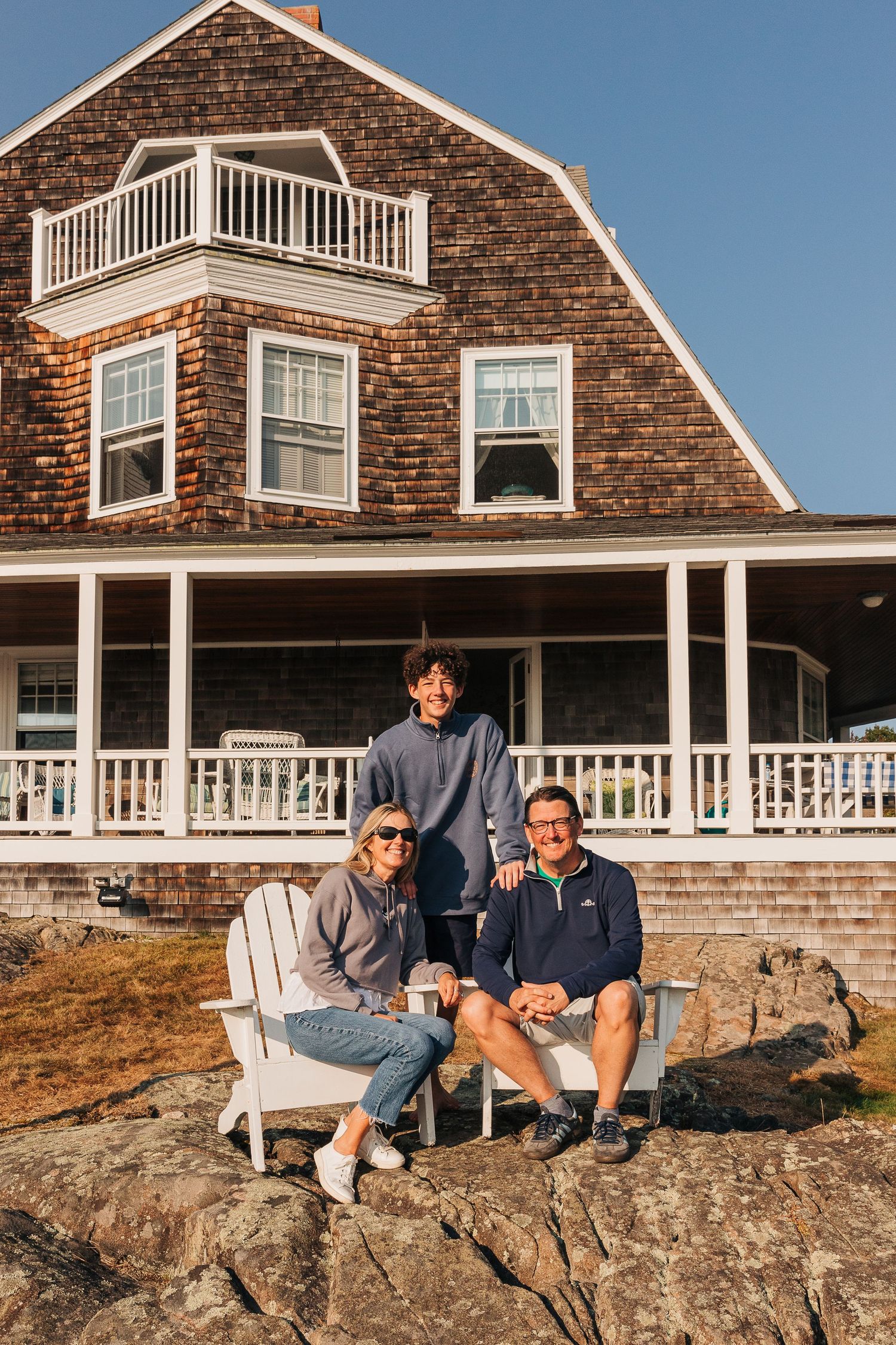Family of 3 sitting on Adirondack chairs in front of a coastal Maine home with wraparound porch.
