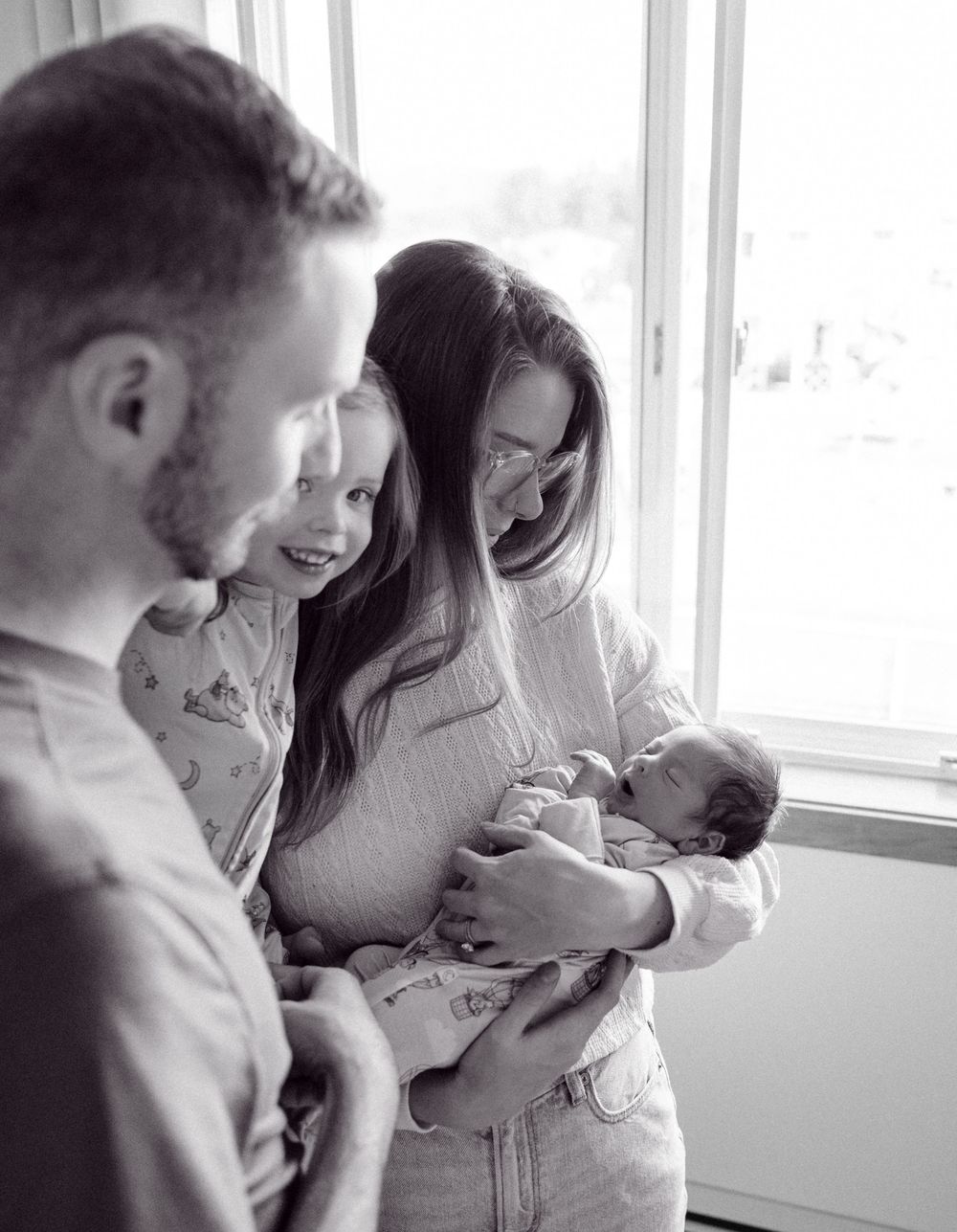 Black and white images of parents sharing intimate moments with their newborn by a bright window.