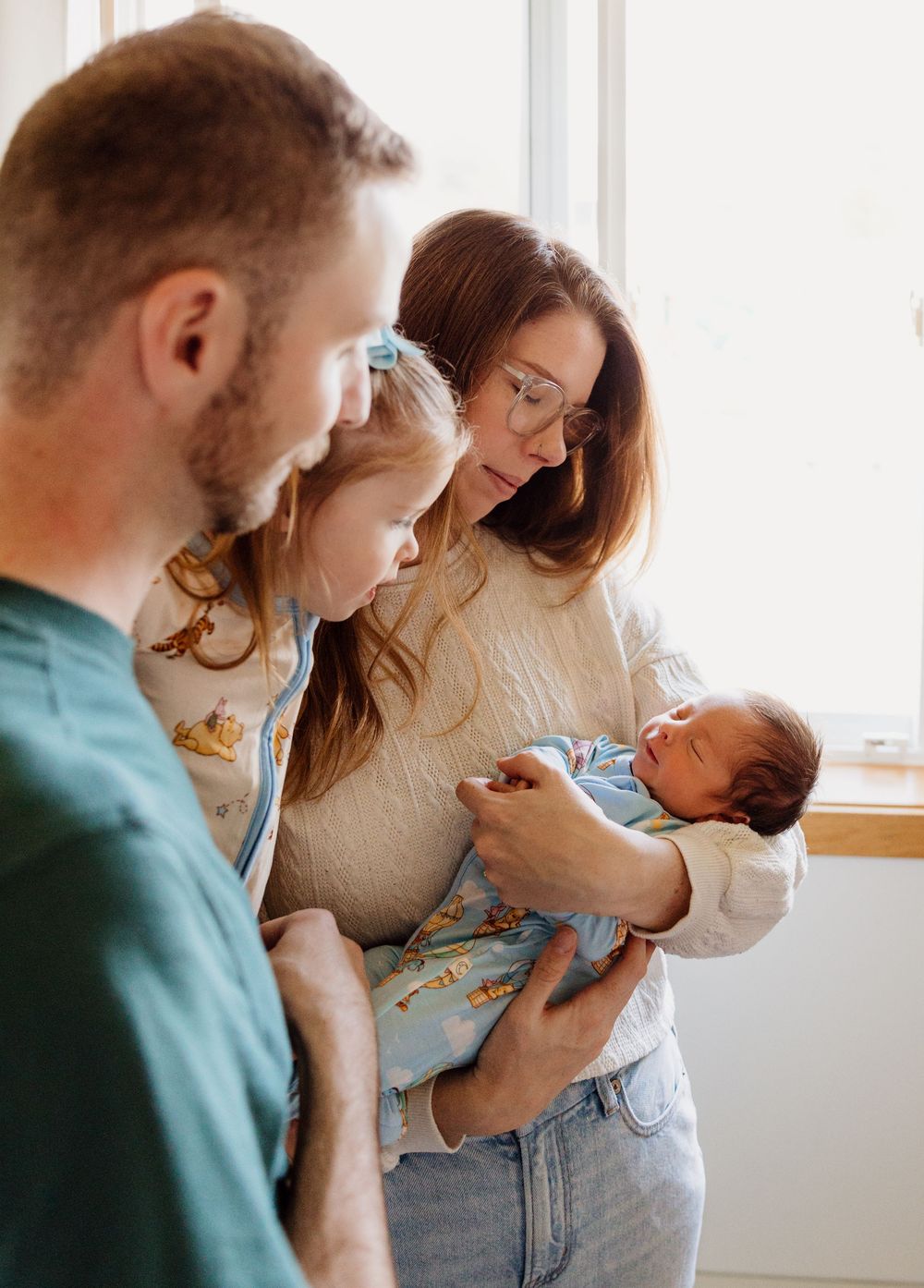 A couple holds and gazes lovingly at their newborn baby in a series of intimate indoor portraits by a window.
