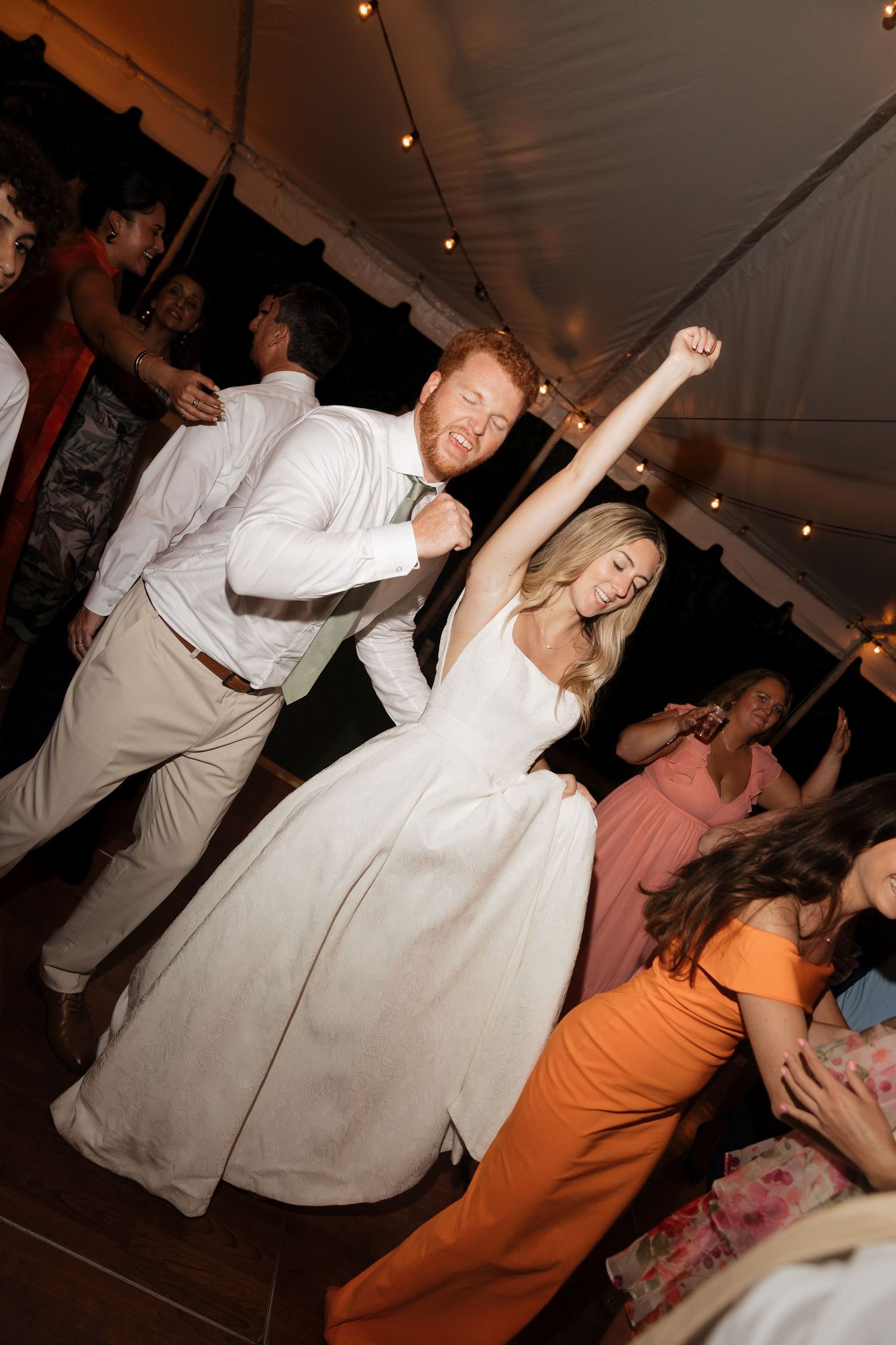 People dancing together at a wedding reception under a tent with string lights overhead.