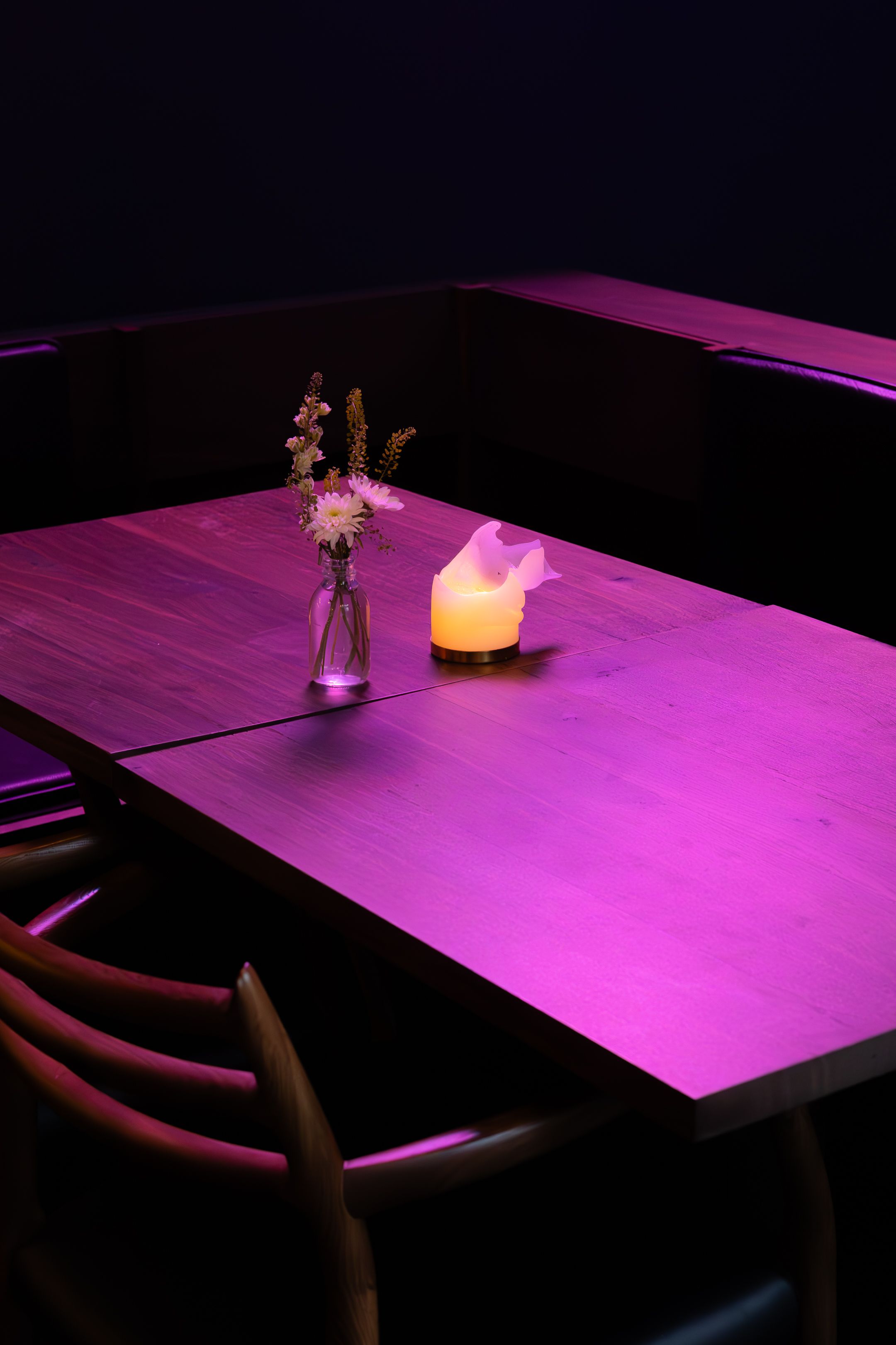 A wooden dining table illuminated by pink ambient lighting with a small vase of flowers and glowing candle centerpiece.