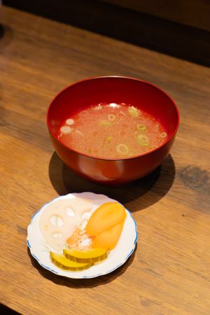 Red ceramic bowl of soup with garnish alongside a small plate on a rustic wooden table.