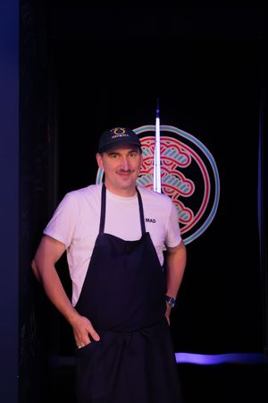 A person in a navy blue apron and white shirt works in a dimly lit restaurant with neon signage visible in background.