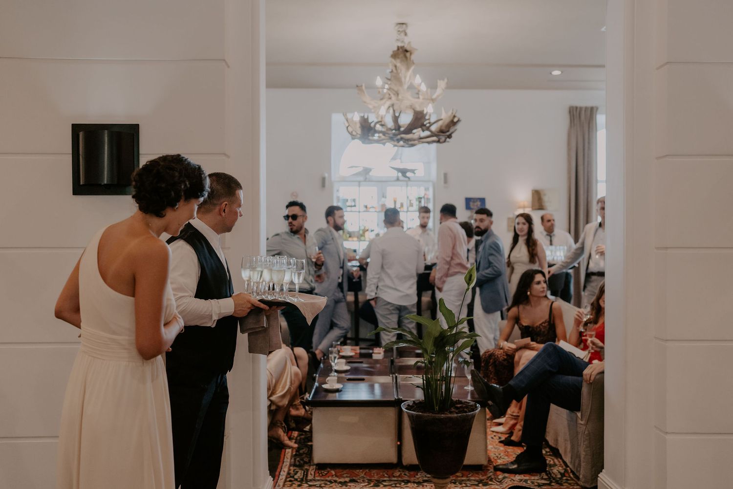 Wedding reception hall with guests gathered around tables and crystal chandeliers overhead.