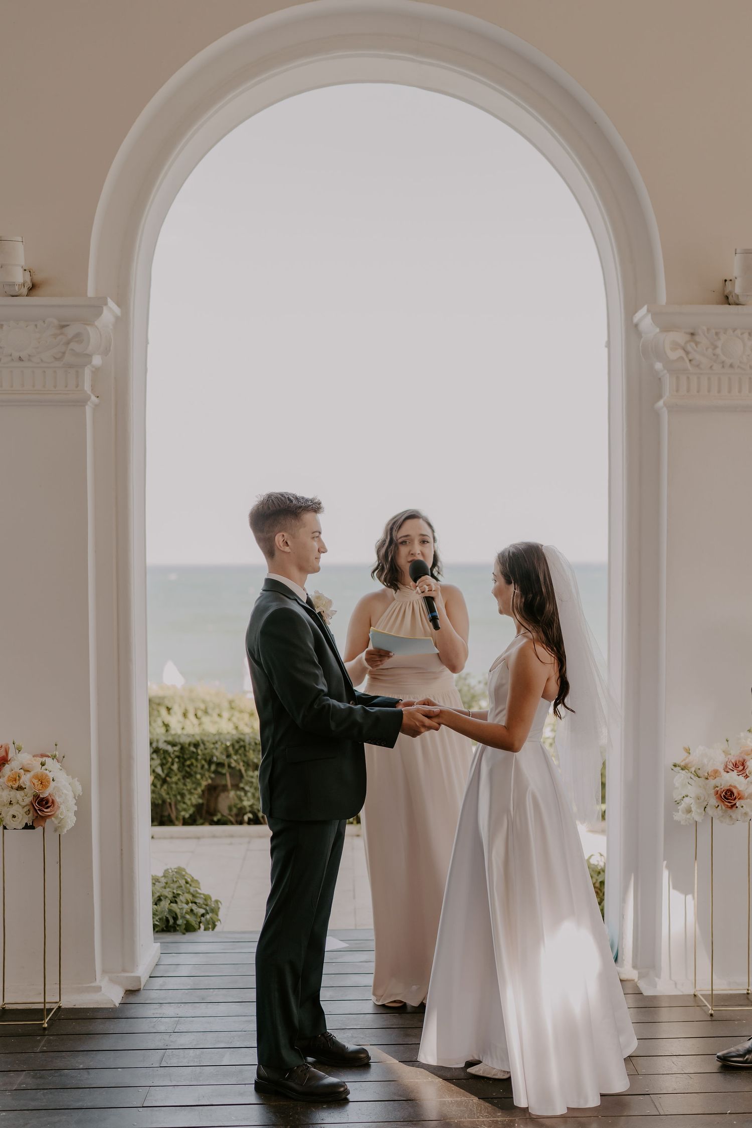 Elegant wedding ceremony taking place under white arched columns with ocean views.
