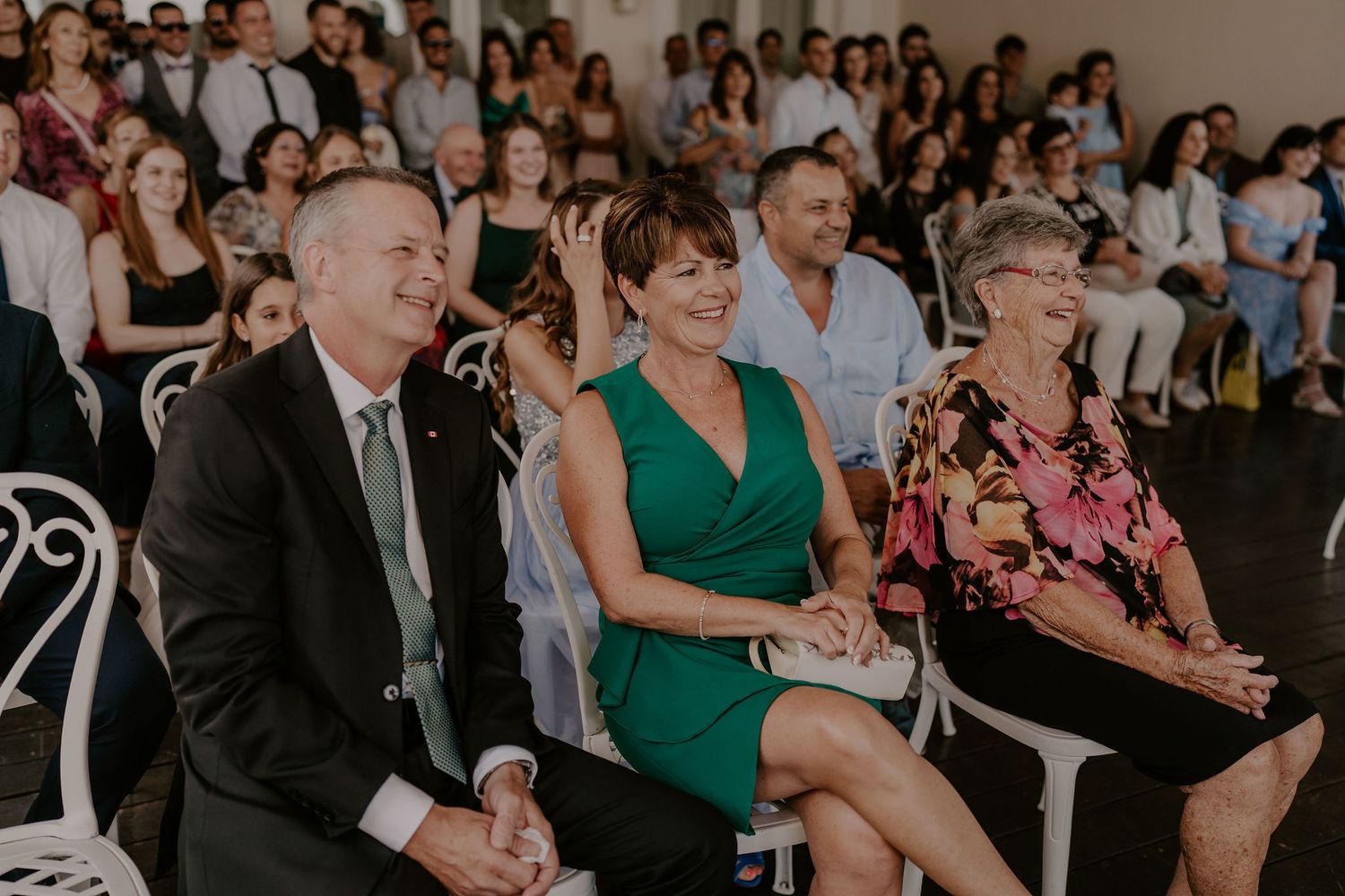 Wedding guests in formal attire smile and laugh while seated in white chairs during an indoor ceremony.