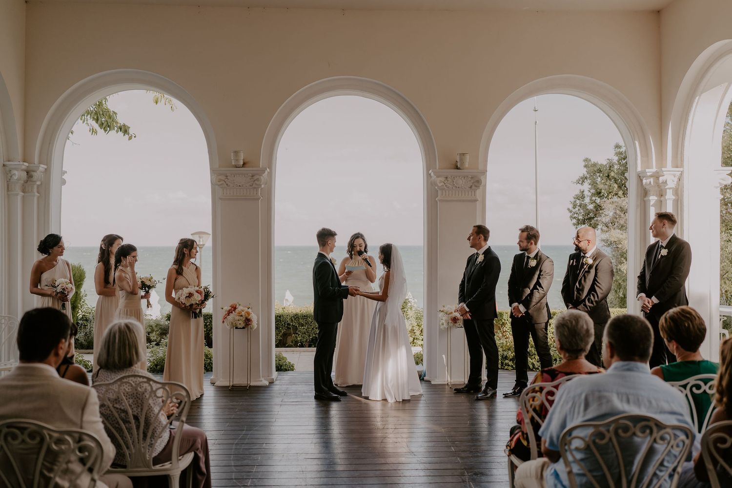 Elegant wedding ceremony taking place under white arches with guests seated in a bright indoor venue.