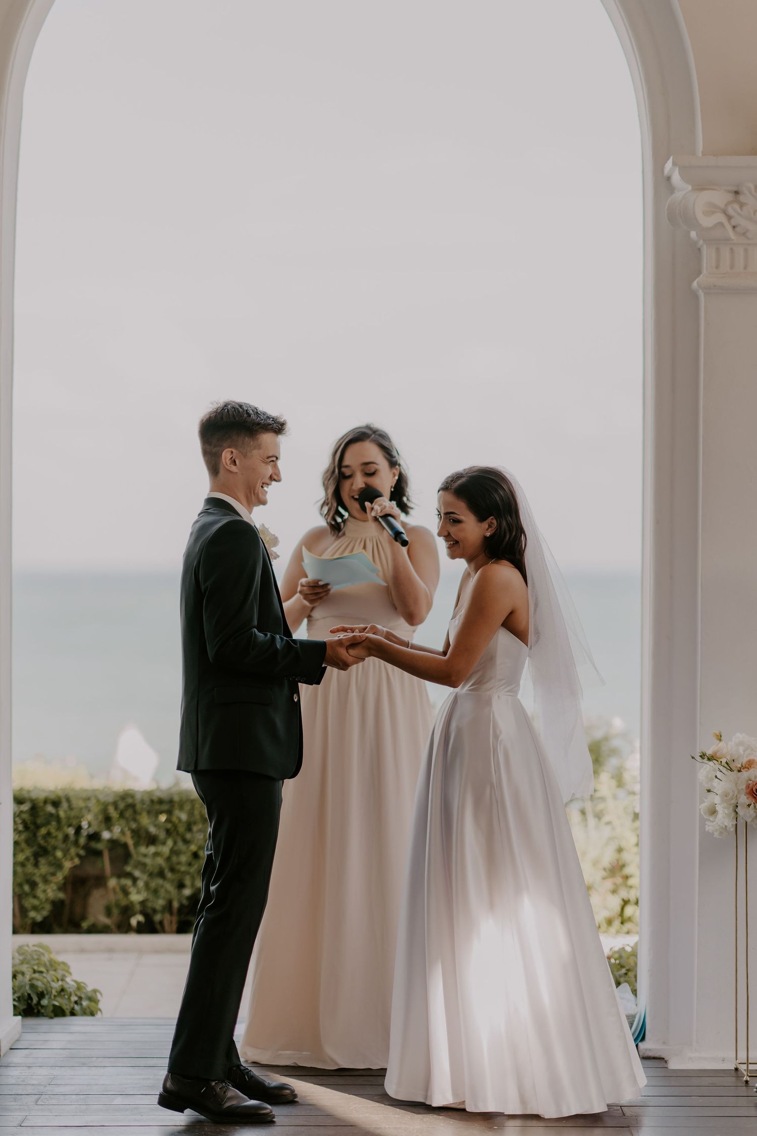 Wedding ceremony under a white archway with officiant and couple exchanging vows by the ocean.