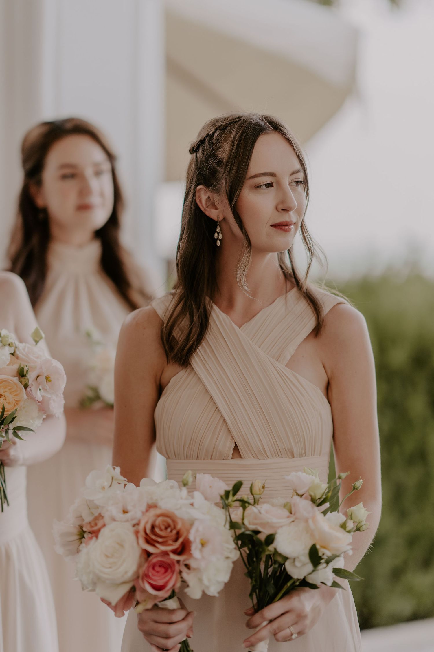 Bridesmaid in champagne crossover dress holds pink and white bouquet during outdoor ceremony.