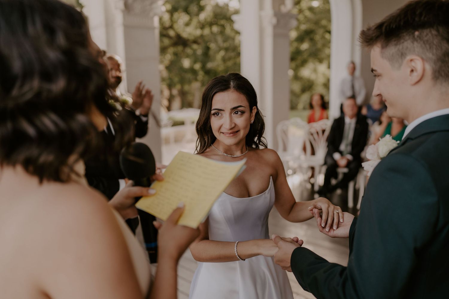 Wedding ceremony participants share a light moment while reading from yellow paper under white arches.