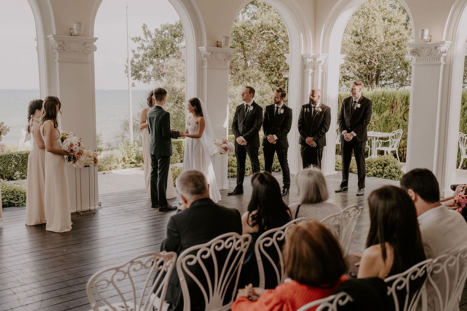 Wedding ceremony in progress under white arched colonnade with guests seated on elegant chairs.