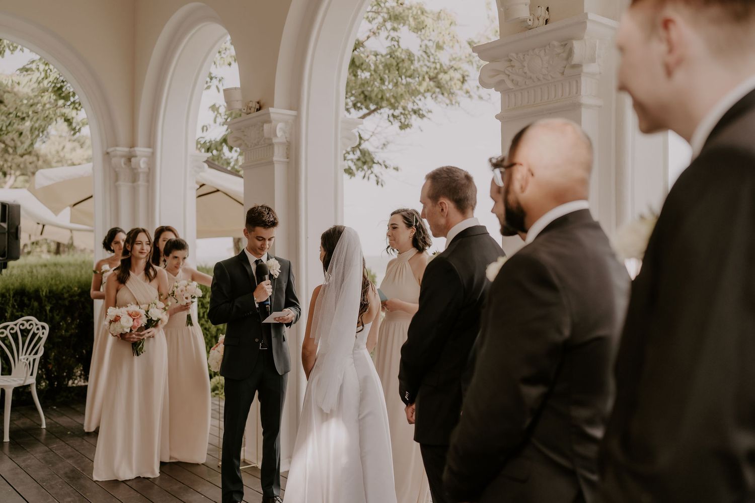 Wedding party gathered under white arched colonnade during outdoor ceremony proceedings.