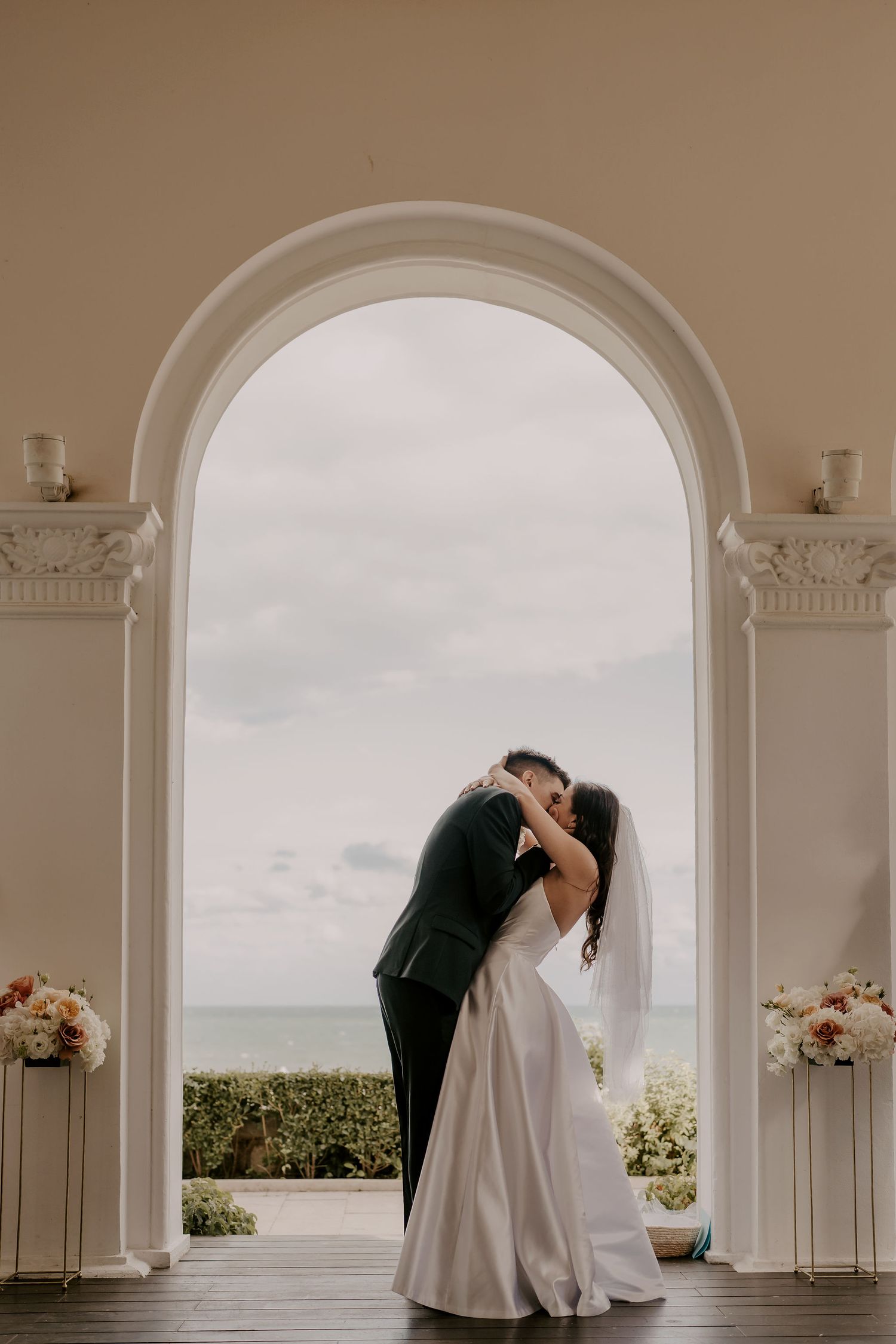 A couple shares their first kiss under an elegant white archway with floral arrangements and ocean views.
