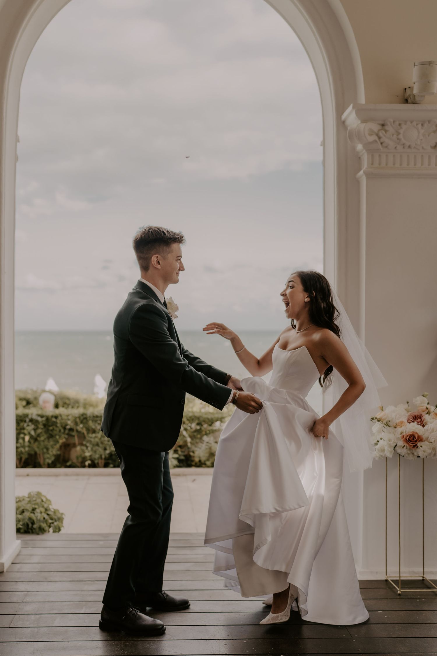 A couple laughs together during a candid moment under a white archway overlooking the ocean.