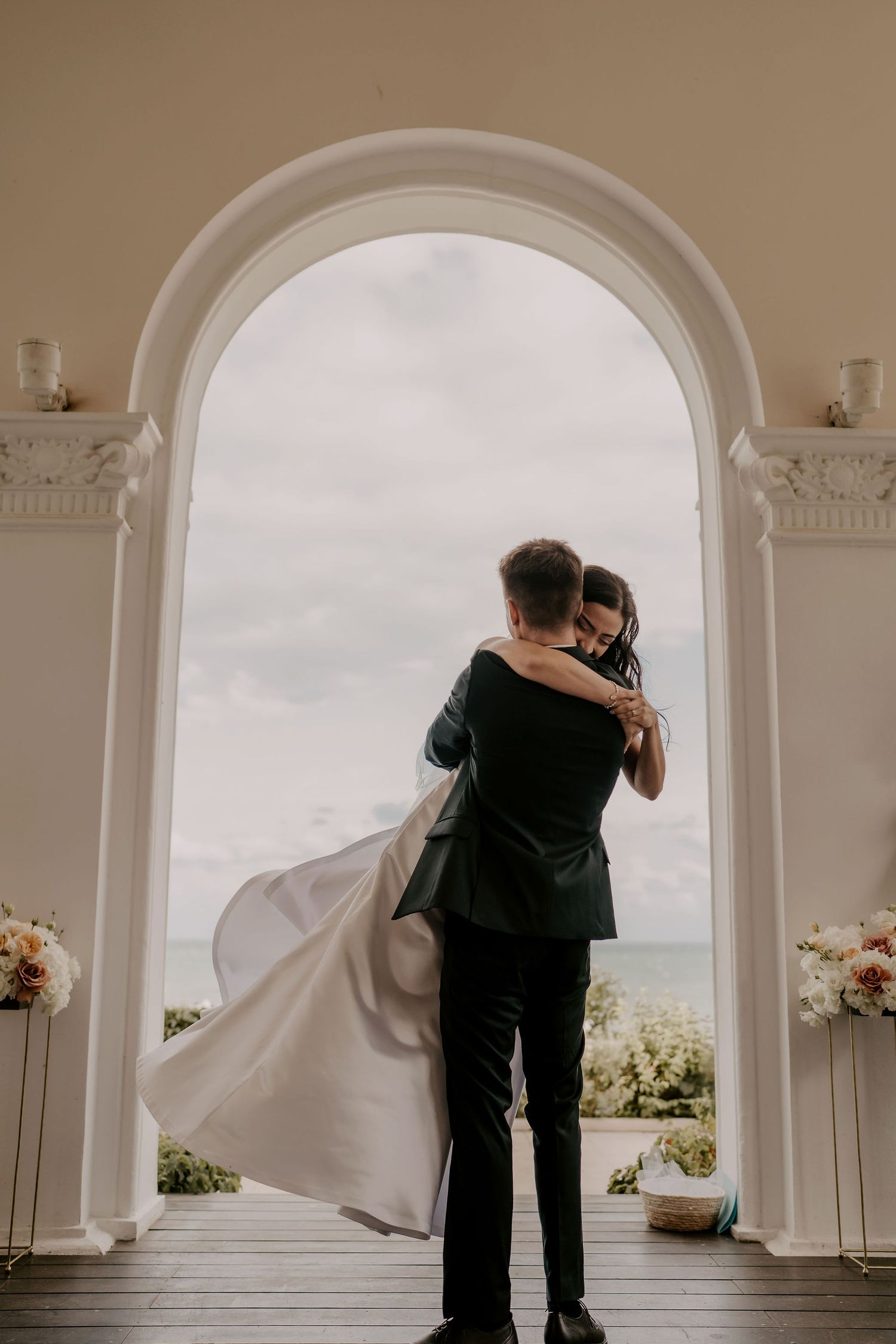 A romantic embrace under an archway with flowing white dress and ocean backdrop.