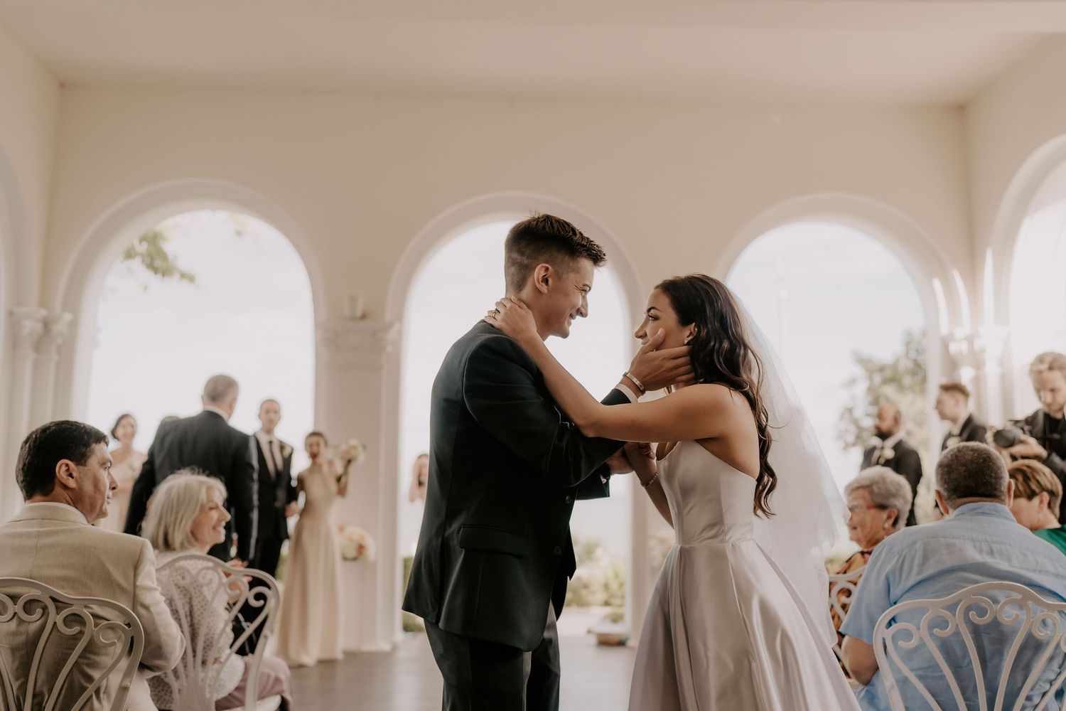 Wedding ceremony in an elegant white room with arched windows and seated guests.